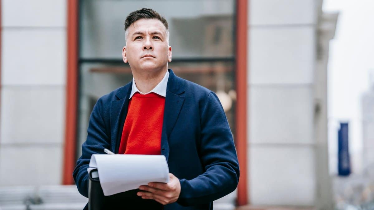 A thoughtful man in professional attire sitting outdoors, looking up pensively.