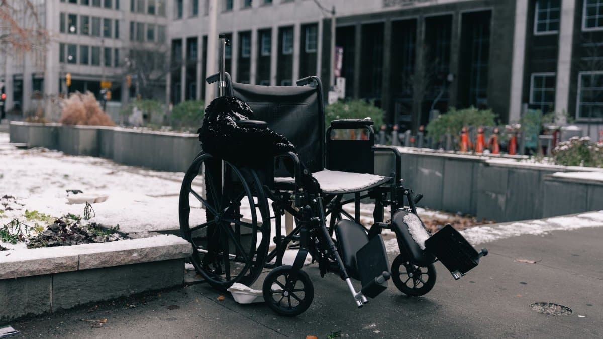 A lone wheelchair covered in snow sits outside in a city, symbolizing neglect and challenges.