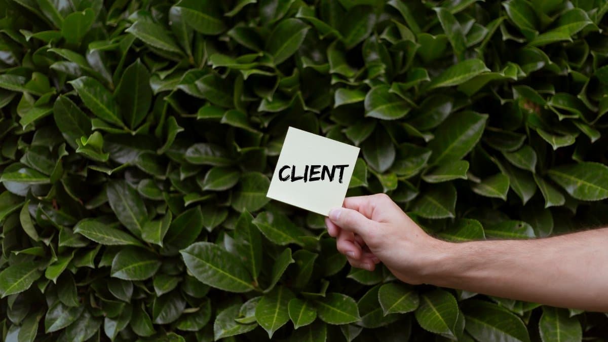 Close-up of a hand holding a note with the word 'CLIENT' in front of a lush green leafy background.