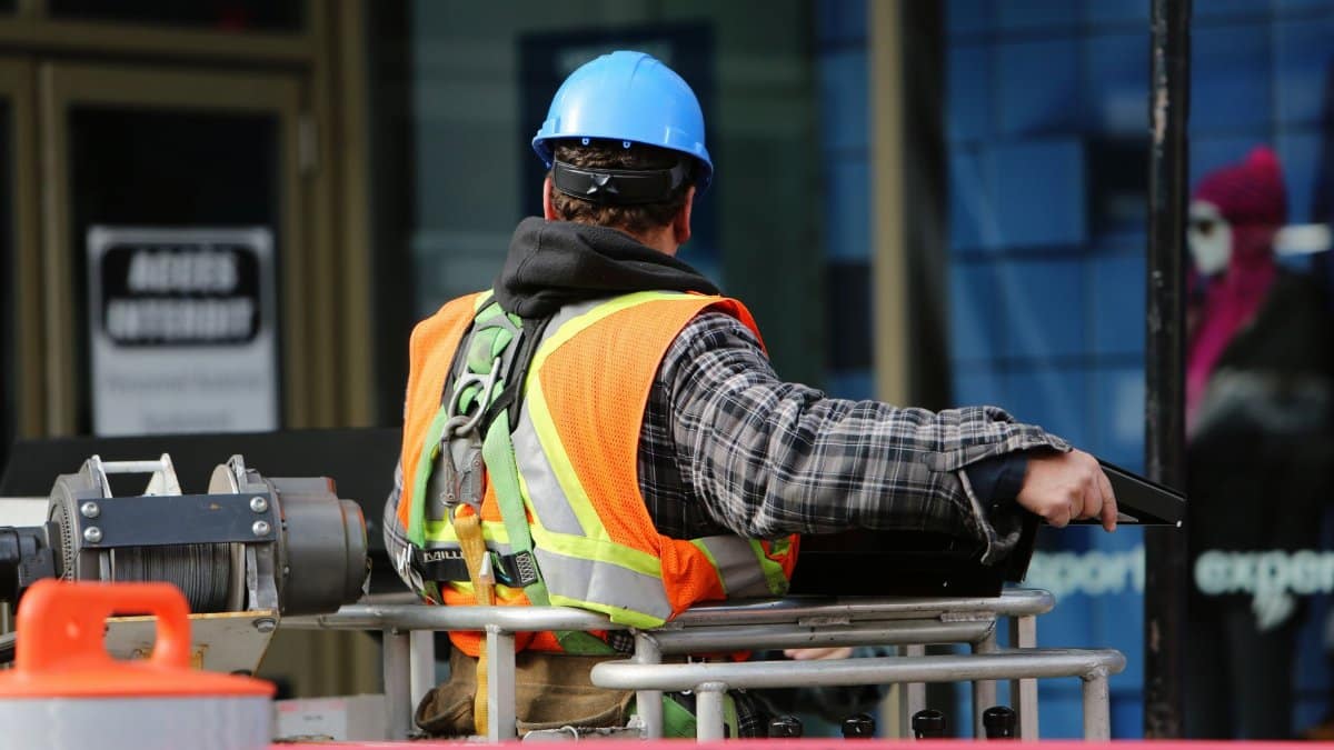 A construction worker wearing a blue helmet and safety vest operates equipment at an urban construction site.