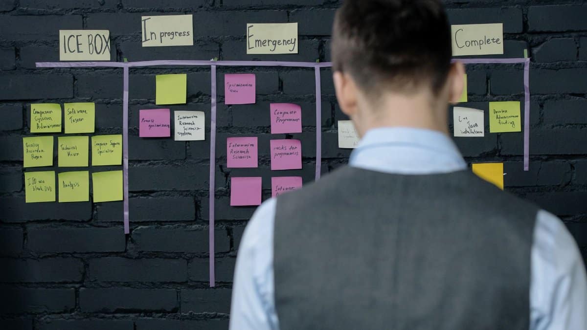 A person in a vest stands in front of a project management board with sticky notes on a dark brick wall.