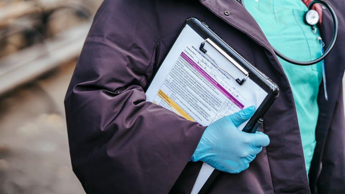 Crop anonymous female doctor in medical clothes and gloves with stethoscope standing outside with documents in clipboard