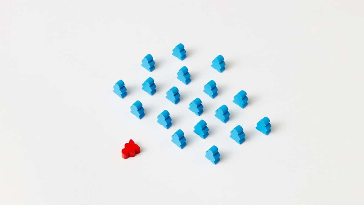 A distinctive red game piece stands out among blue meeples on a minimalist white background, symbolizing individuality.