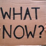 Cardboard sign reading 'What Now?' held outdoors, conveying uncertainty or protest.