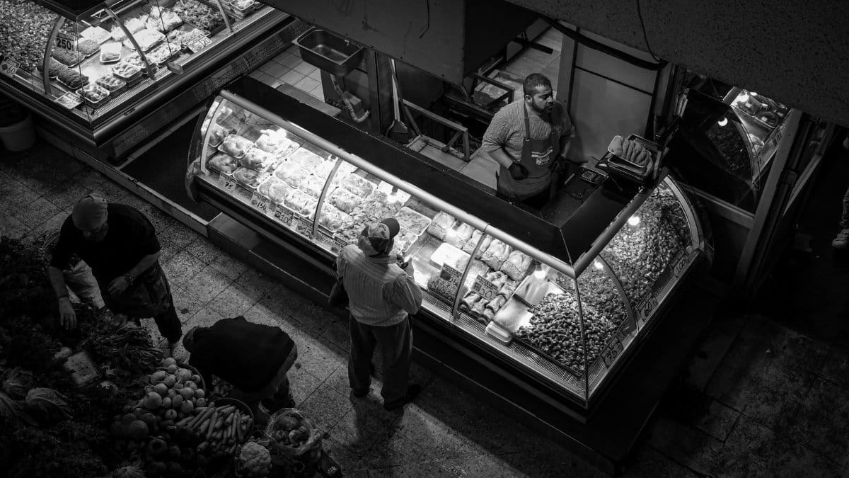 A vibrant market scene with a vendor engaging customers at a deli counter, captured in black and white.