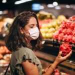 Woman wearing a mask while selecting fresh dragon fruit at a supermarket fruit aisle.
