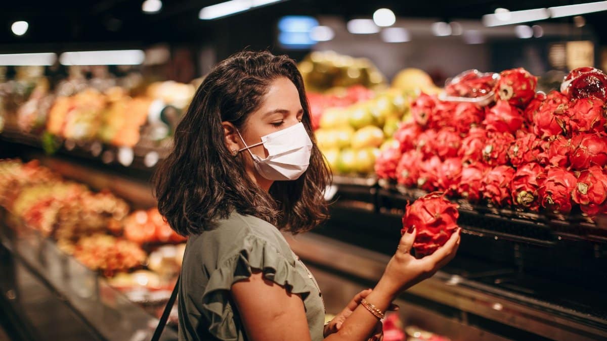 Woman wearing a mask while selecting fresh dragon fruit at a supermarket fruit aisle.