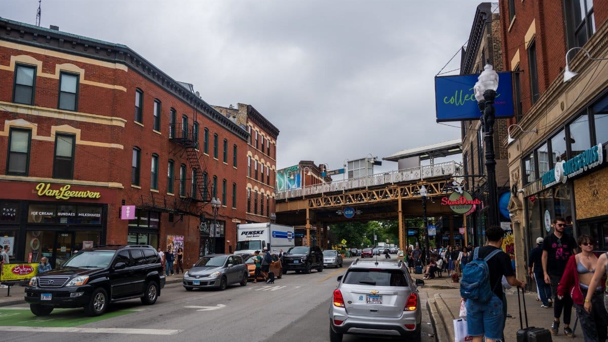 Vibrant Chicago street scene with shops, cars, pedestrians, and elevated train track overhead.