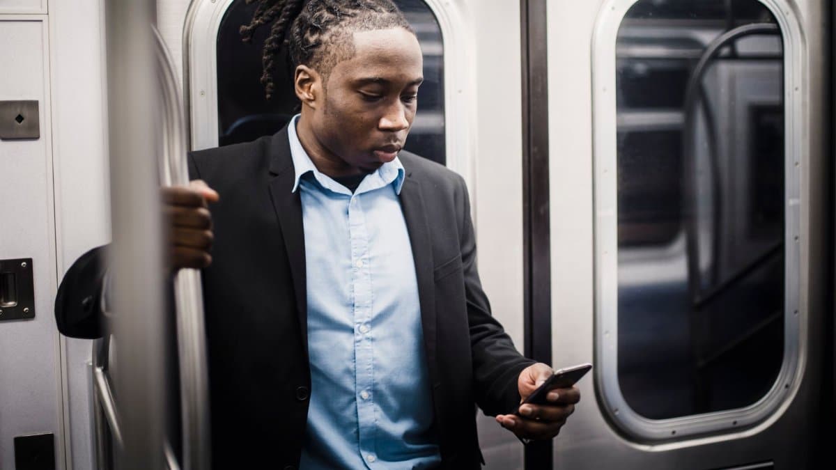 Adult well dressed African American male worker surfing internet on mobile phone and holding handrail while commuting to work on subway