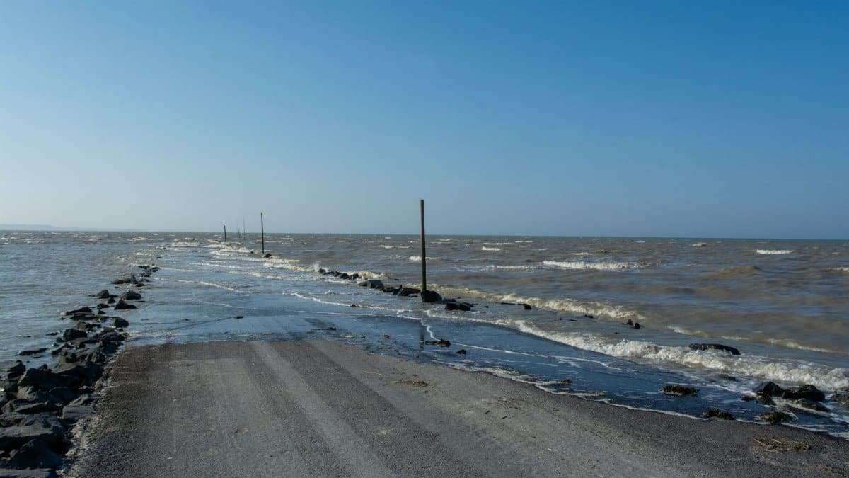 A flooded coastal road surrounded by the sea and horizon under a clear blue sky.