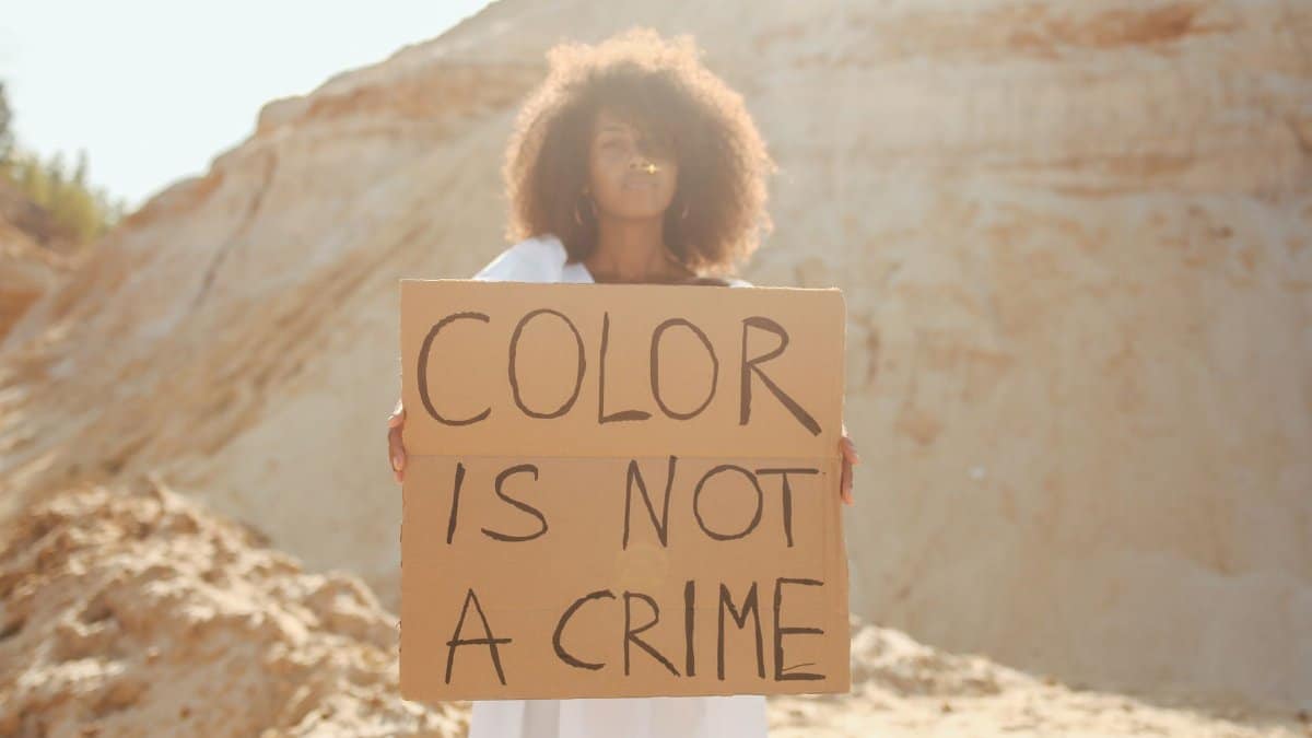 Woman holding a sign reading 'Color Is Not a Crime' in a sandy outdoor setting.