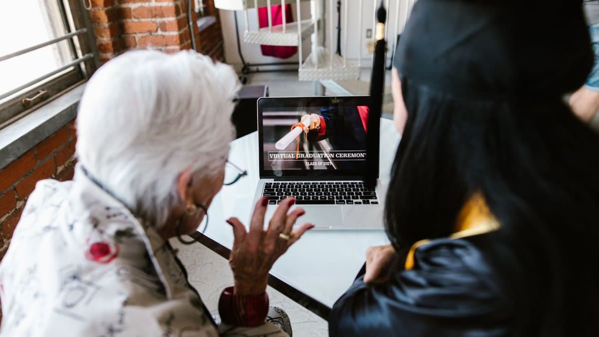 A senior and a graduate watch a virtual graduation ceremony on a laptop at home.