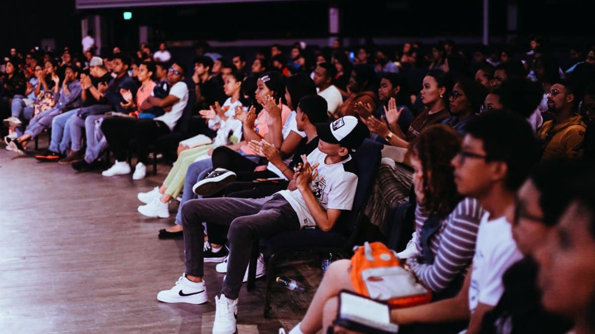 A diverse crowd seated in an auditorium applauds a live performance.