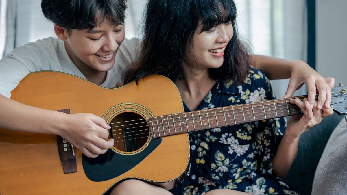 A joyful lesbian couple enjoys playing an acoustic guitar together indoors, showcasing their close relationship and shared passion for music.