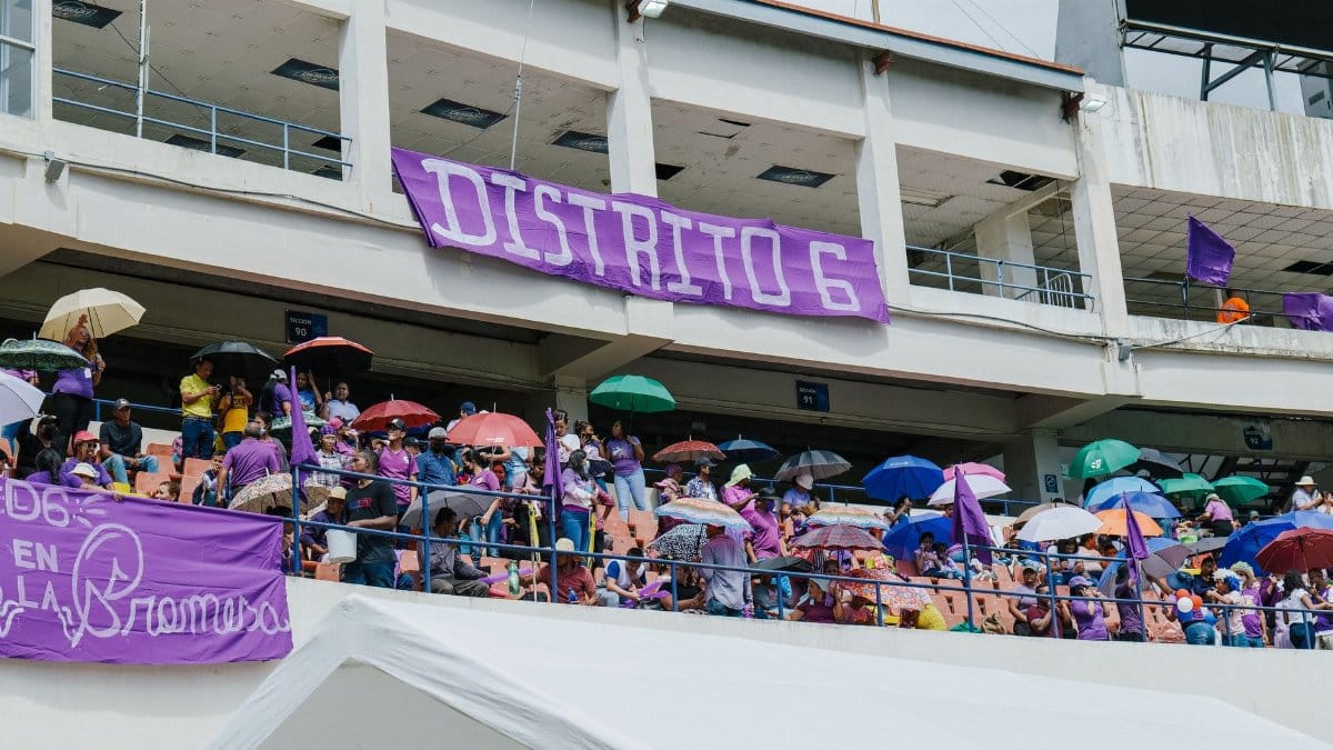 A lively crowd in a stadium stands under umbrellas during a community event on a bright day.