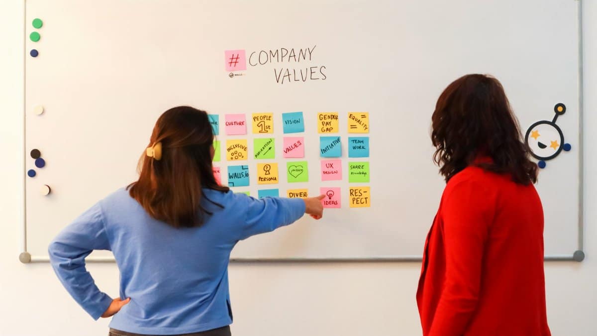 Two women discussing company values on a whiteboard in an office setting, emphasizing teamwork.