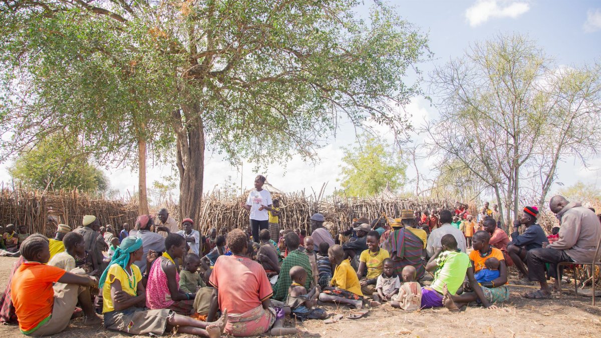 A diverse group of adults and children gather under a tree for a community meeting outdoors.