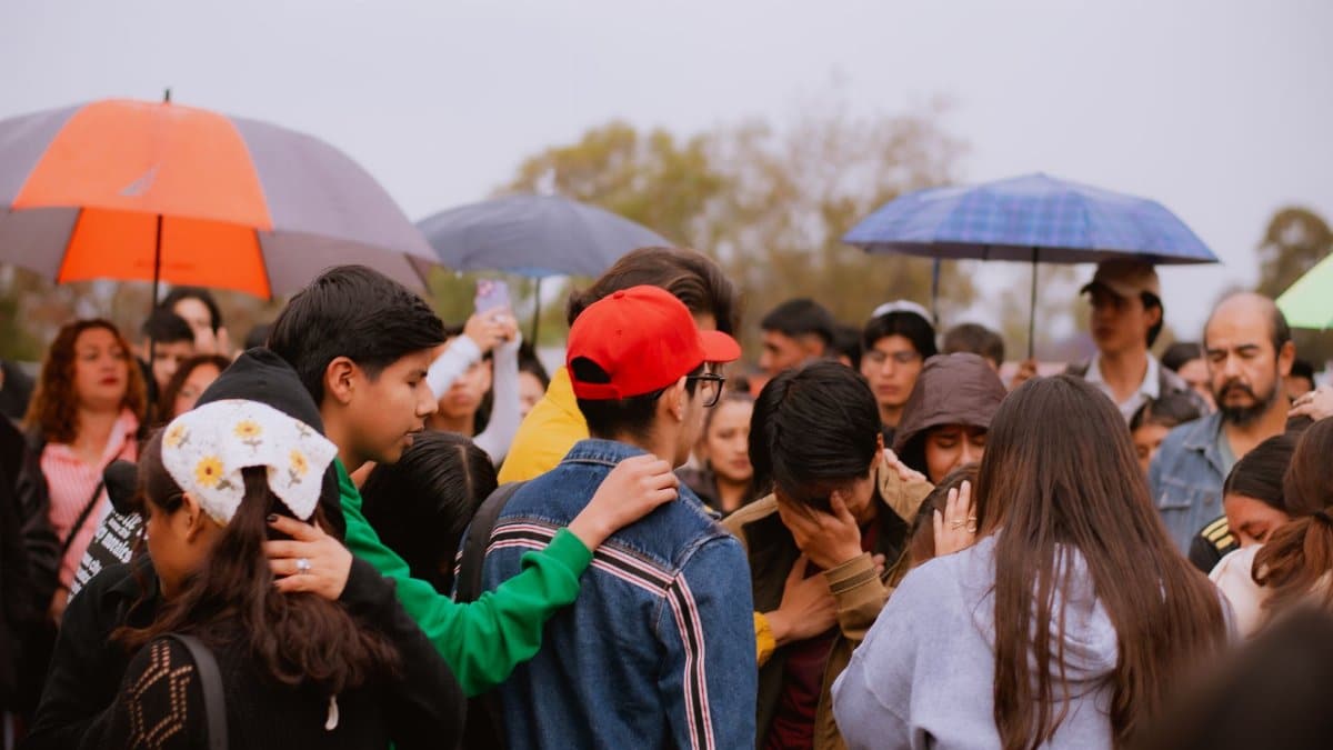A group of diverse adults and teenagers gather under umbrellas on a cloudy day, showing support and empathy.