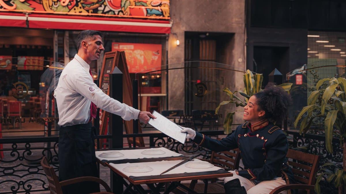 A waiter serves a menu to a seated customer at an outdoor restaurant with colorful signage.