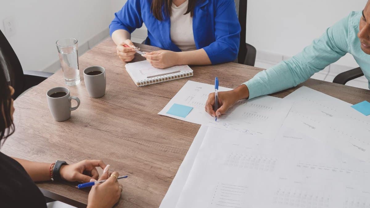 A group of adults in a business meeting, discussing plans around a table with documents.