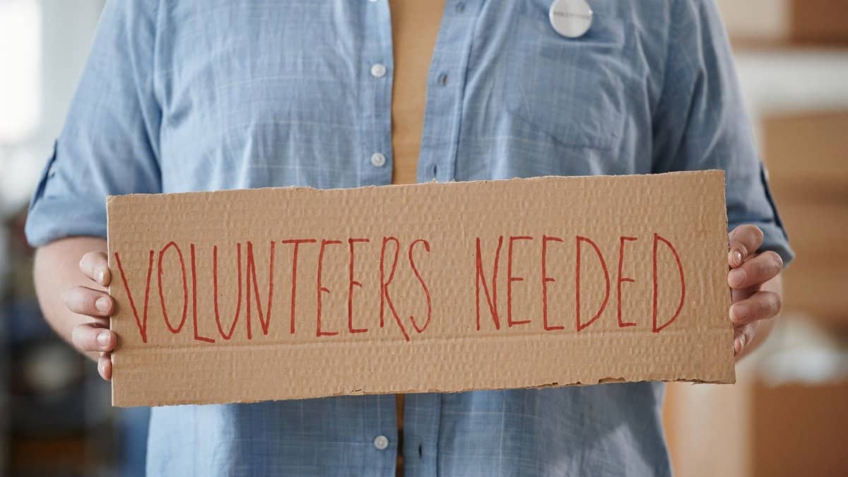 Person holding a sign reading 'Volunteers Needed' to encourage community support.