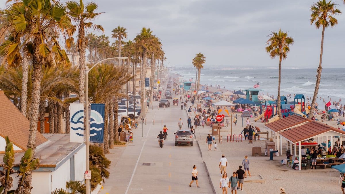 Bustling beach and boardwalk with palm trees, people, and ocean views during sunset.