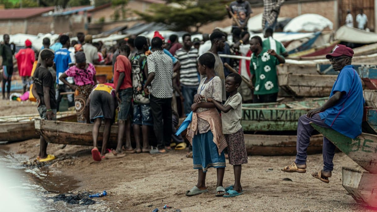 A bustling scene of a community gathering near wooden boats along a waterfront.
