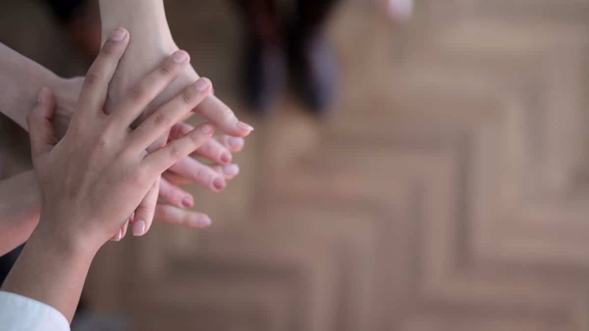 Unrecognizable crop people holding hands together in teamwork and support above wooden brown floor
