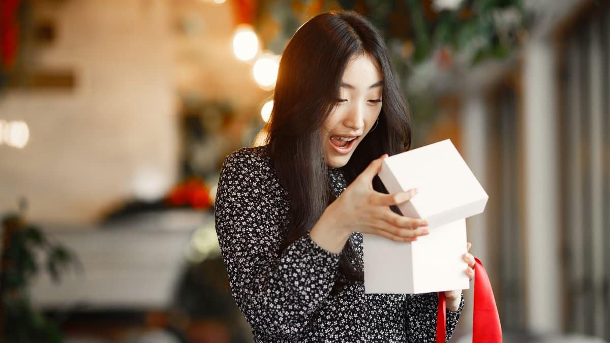An Asian woman delightfully opens a gift box with a red ribbon indoors.