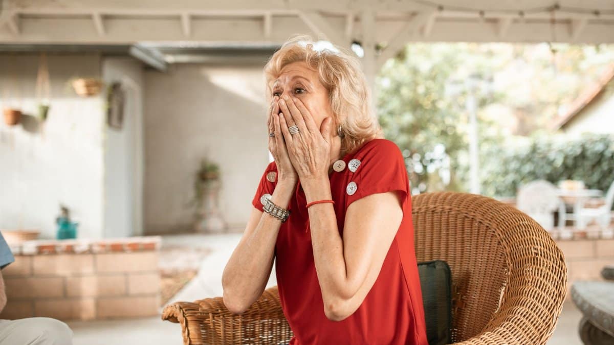 An elderly woman with a surprised expression sits in a wicker armchair indoors.