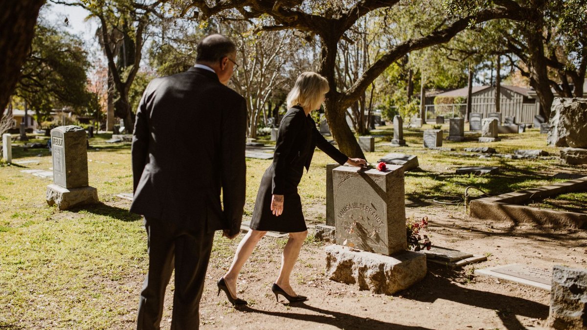 A somber moment as a couple places a rose on a gravestone in a peaceful cemetery.
