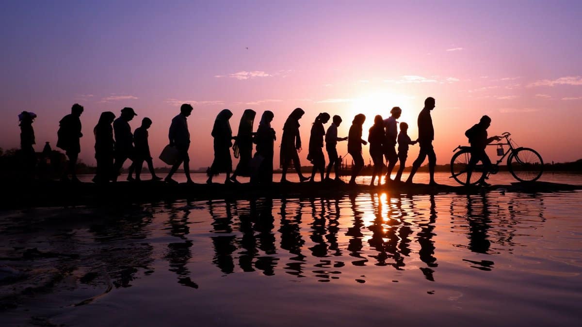 A line of silhouetted people walking along a waterfront at sunset with reflections on the water.