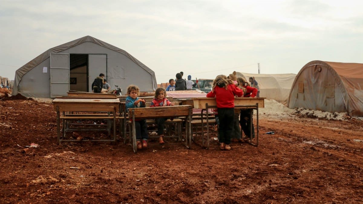Children gather around desks in a refugee camp in Idlib, Syria, highlighting resilience and community.