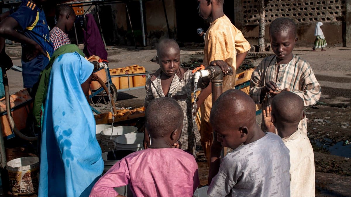 A group of children collect water from a communal tap in a rural setting, highlighting daily life.