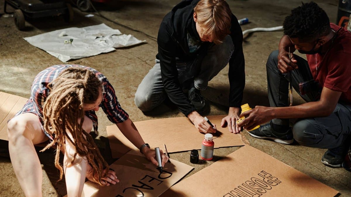 Three adults making protest signs with cardboard and markers indoors.