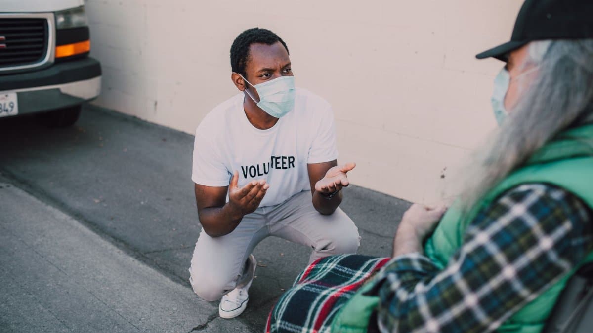 A compassionate volunteer wearing a face mask interacts with a community member outdoors, symbolizing social good.