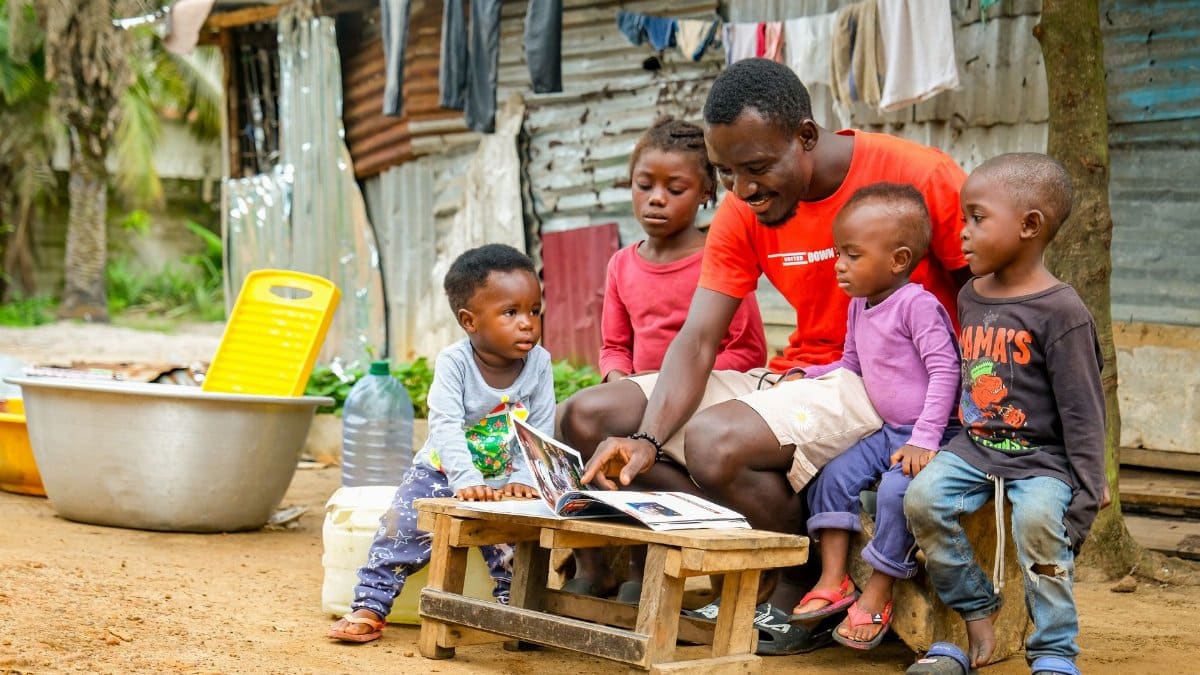 A father reads a book to young children outside a tin house in Monrovia, fostering learning and community.