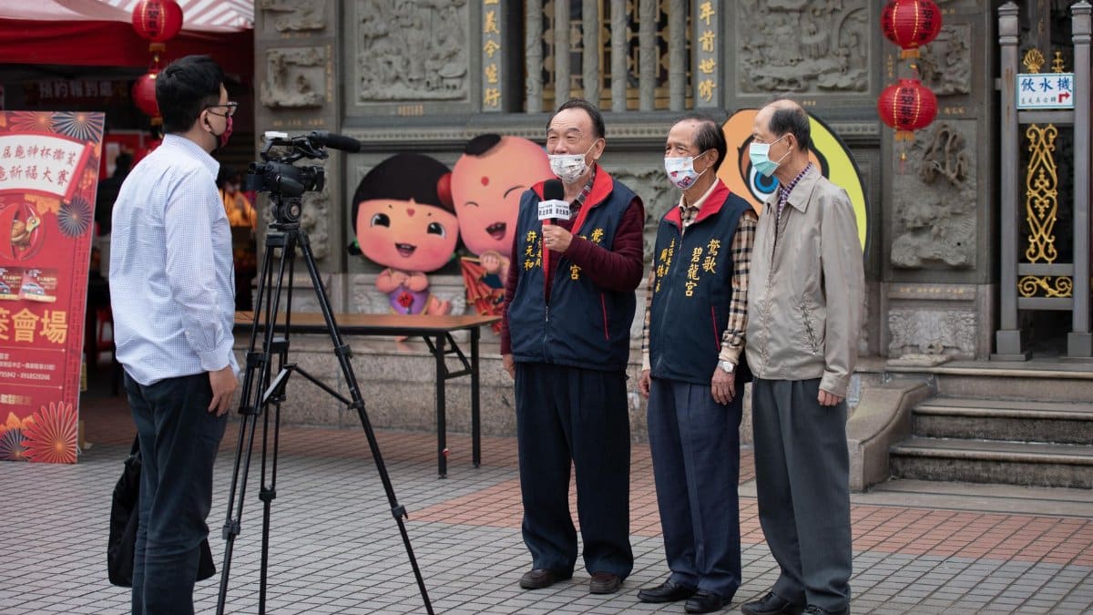 Elderly men wearing face masks being interviewed outdoors with festive decorations in the background.