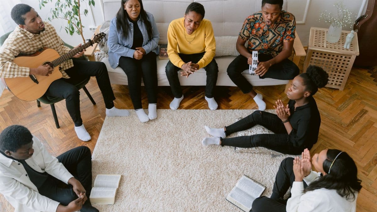A diverse group of people praying, singing, and playing guitar indoors, emphasizing faith and community.