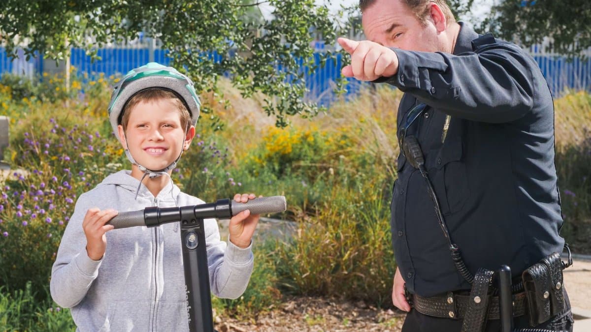 A police officer assists a young boy on a scooter at a park, signifying community safety.