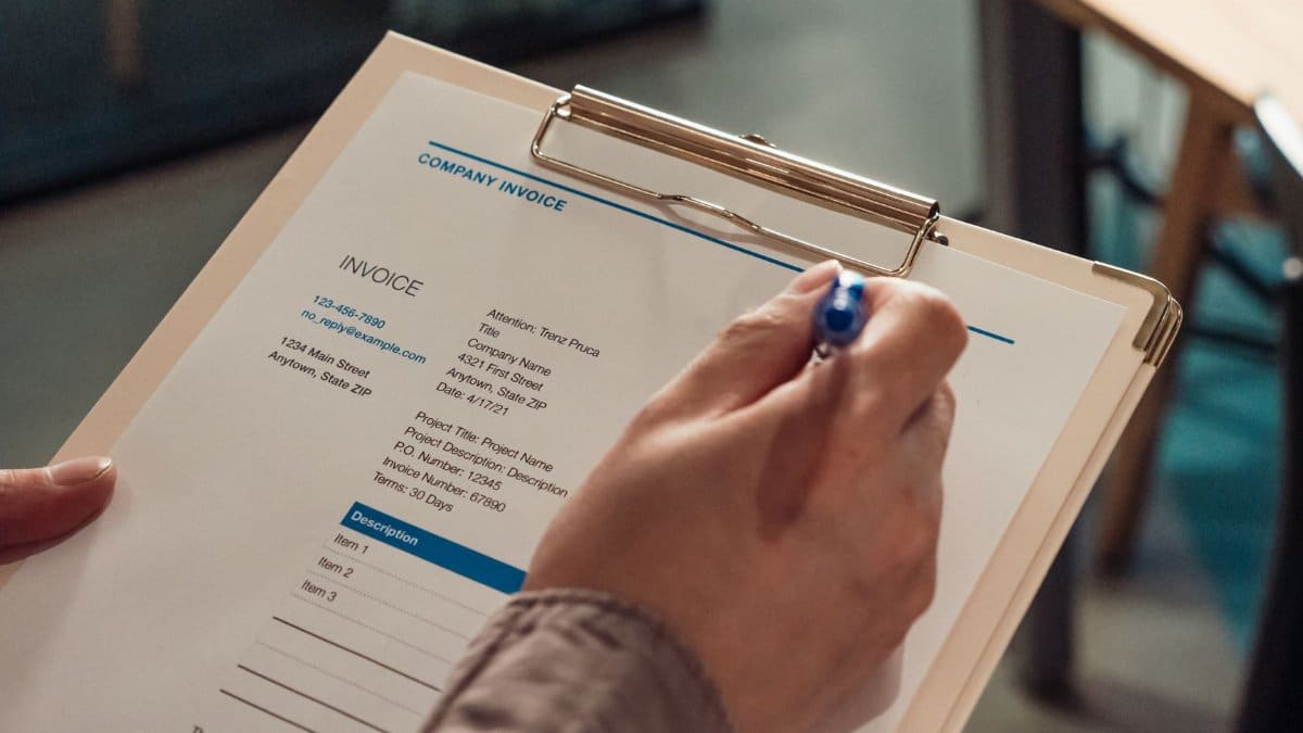 Person's hand holding a company invoice on a clipboard with a pen.