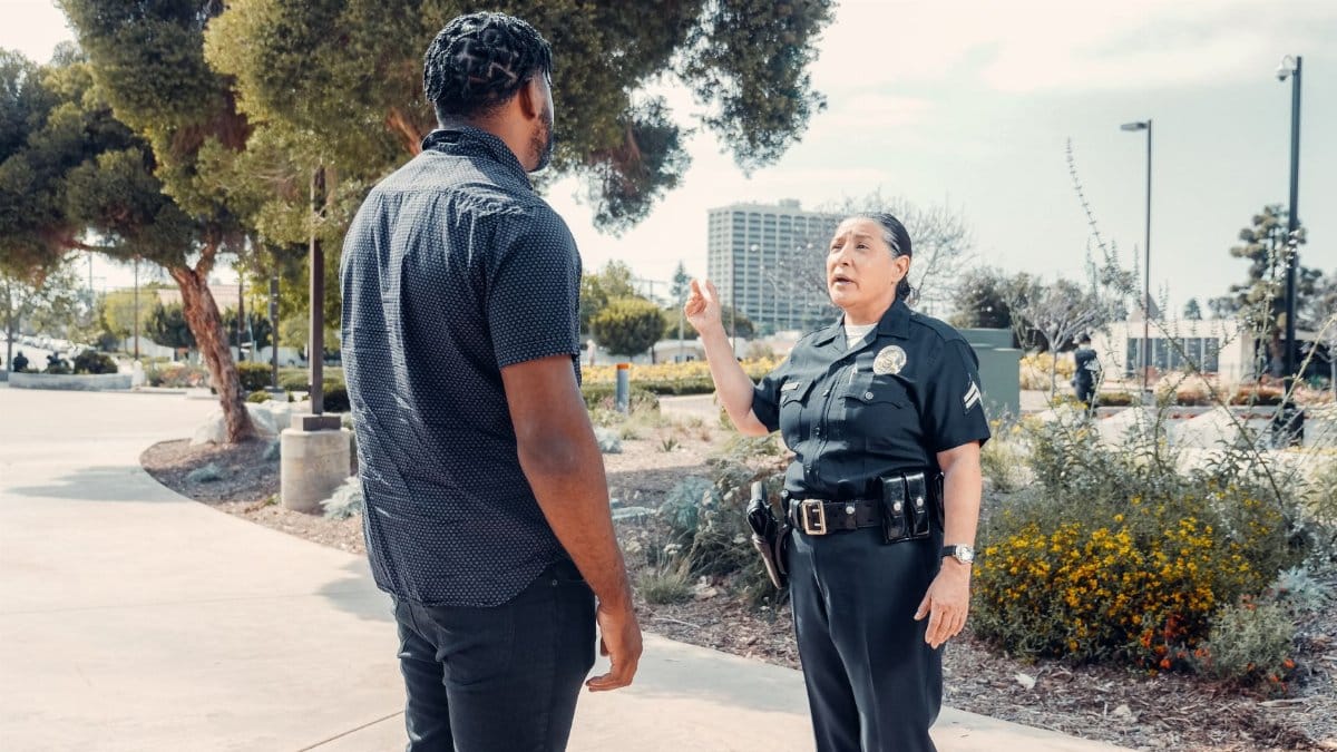 A police officer engages in conversation with a civilian in a park setting on a sunny day.