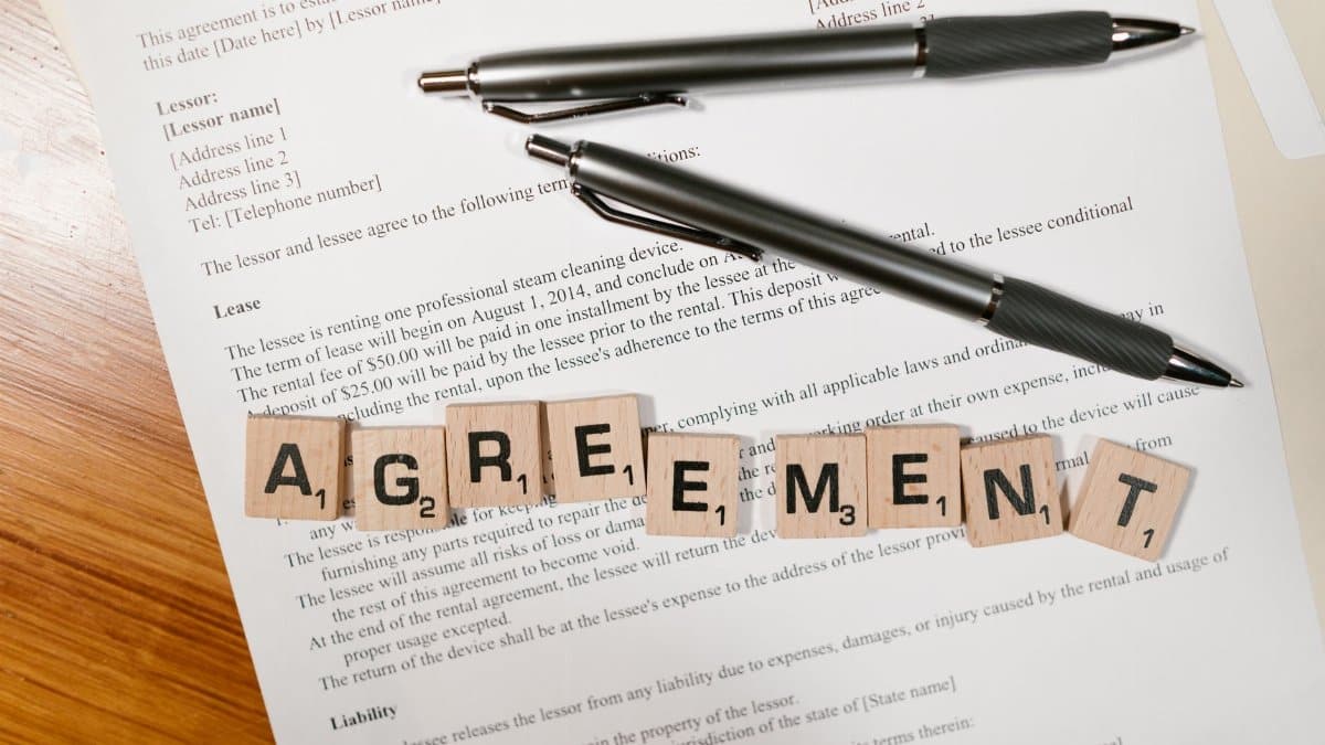 A document with Scrabble tiles spelling 'AGREEMENT' and two pens, symbolizing contract signing.