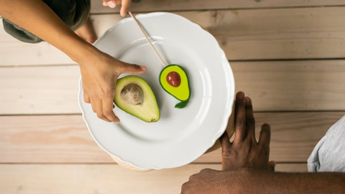 Top view of crop anonymous African American people near white plate with half of ripe avocado and sweet lollipop with similar shape