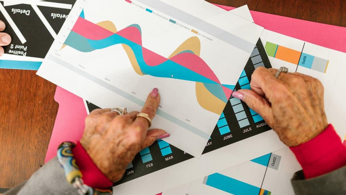 Senior adult pointing at colorful business graphs with data analysis documents on a wooden table.