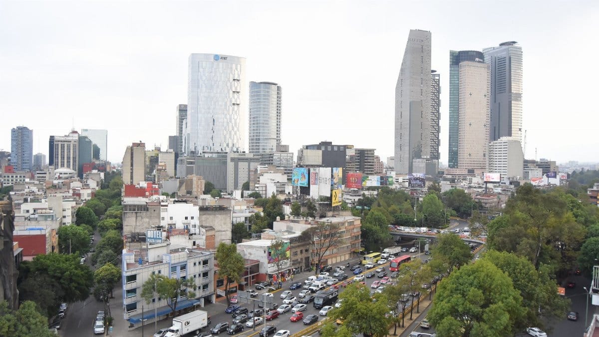 A bustling Mexico City street with skyscrapers, traffic, and greenery under a cloudy sky.