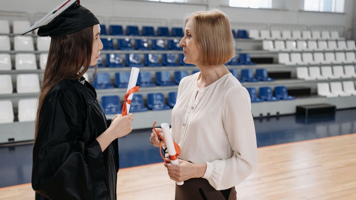 Two women discussing during a graduation ceremony, each holding diplomas.