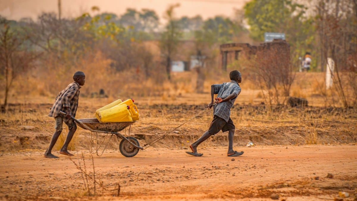 Two children transporting water containers on a wheelbarrow in rural Adjumani, Uganda.