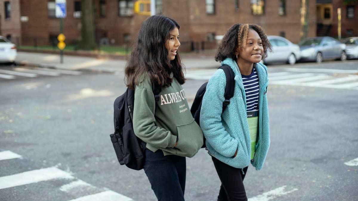 Content multiracial girlfriends with school bags and hands in pockets interacting on urban crosswalk while looking away
