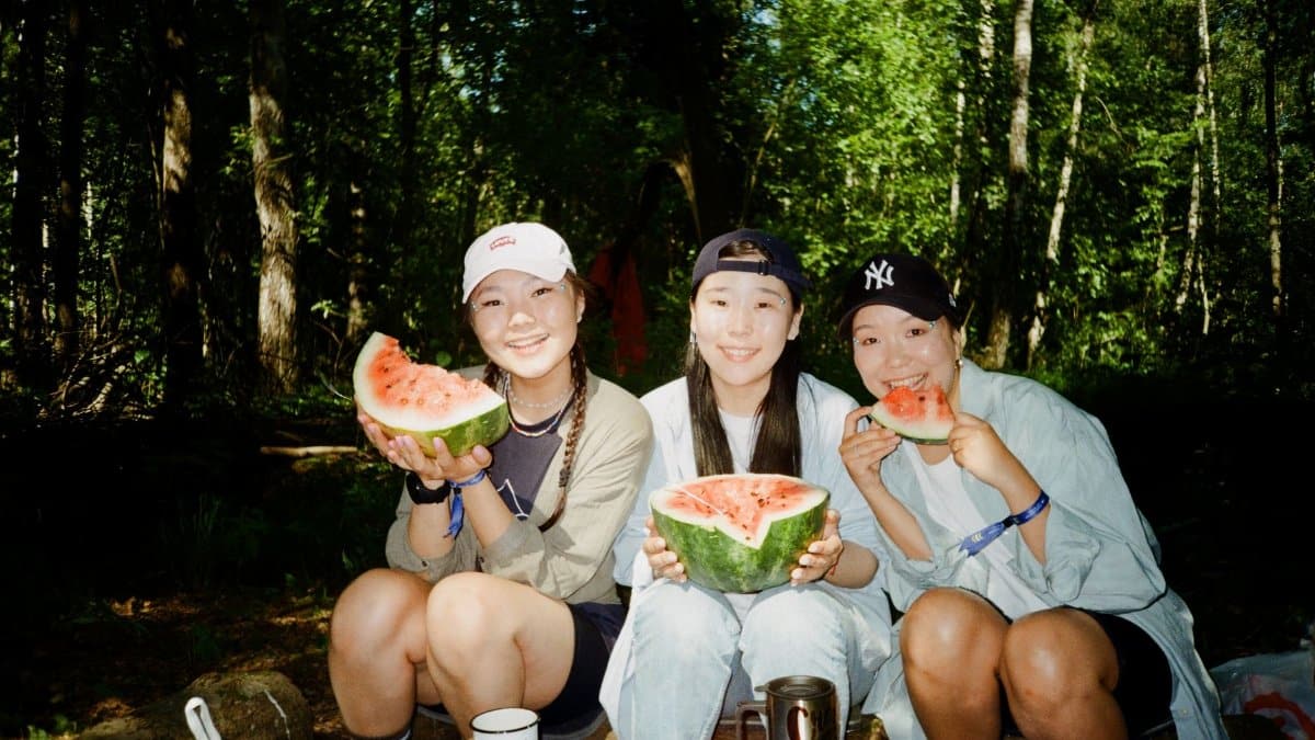 Three friends happily eating watermelon together in a summer forest setting.